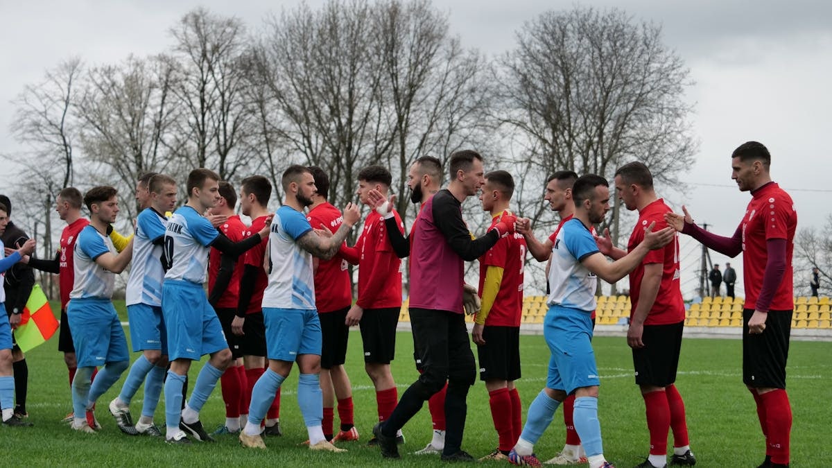 Football teams in red and blue kit shaking hands on a grassy pitch before the game.